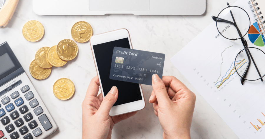 Person holding a credit card and a smartphone, with a calculator, gold coins, glasses, and financial charts on the table, ready to automate payments.